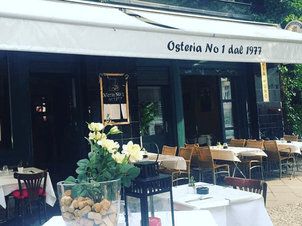 Die Fassade der Osteria N.1 bei Tag: weiße Markise mit der Aufschrift Osteria No 1 dal 1977, davor gedeckte Tische
